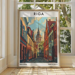 Op de afbeelding: Een ingelijste kunstdruk met een kleurrijke illustratie van een straat in Riga met kasseien, historische gebouwen en een centrale kerktoren. De tekst RIGA staat bovenaan, met RIGA, LATVIA en 56.95° N, 24.11° E onderaan.