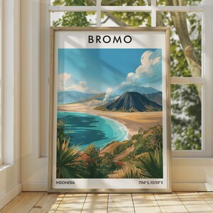 Puede incluir: Un póster vertical enmarcado que presenta una ilustración estilizada del Monte Bromo en Indonesia. La obra muestra un paisaje volcánico con una montaña oscura, una llanura arenosa, agua azul y vegetación verde. El texto BROMO, INDONESIA, 7.94 S, 112.95 E aparece.