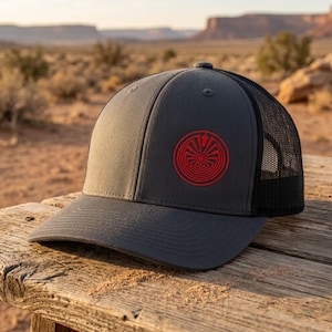 May include: A charcoal grey baseball cap with a black mesh back and a red circular graphic featuring a labyrinth design on the front panel. The cap has a structured crown, a curved brim, and a solid grey front section with visible stitching details.