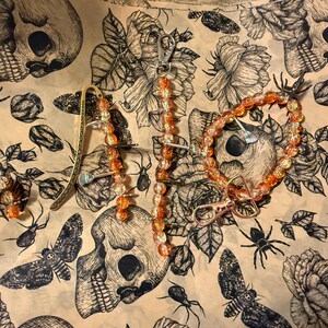 May include: An assortment of orange beaded jewelry, including a bracelet, earrings, and a keychain, is displayed on a beige fabric with a black and white skull and floral pattern. The jewelry features clear and orange beads.