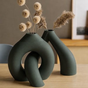Puede incluir: Dos jarrones de cerámica de color verde oscuro con acabado mate. Uno presenta un diseño circular de bucle anudado, mientras que el otro tiene una forma tubular alta y curva. Ambos jarrones contienen tallos botánicos secos de color beige.