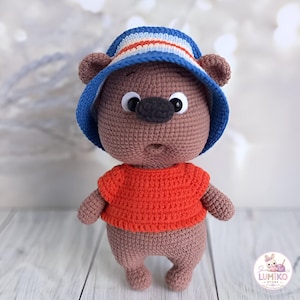 Può includere: Una bambola a forma di orso lavorata all'uncinetto di colore marrone, che indossa una maglietta arancione brillante e un cappello da pescatore a righe blu, bianche e arancioni. L'orso ha occhi in plastica nera, naso grigio scuro e bocca aperta.