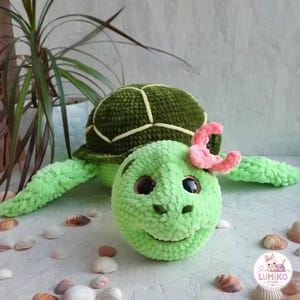 May include: A plush crochet turtle toy made of soft light green yarn with a dark olive green shell featuring light yellow stitching. The turtle has large dark brown eyes and a small pink crocheted flower attached to the side of its head.