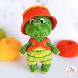 Op de afbeelding: Een gehaakte schildpaddenpop met een groene kop en lichaam met lichtgroene strepen, met een oranje vissershoedje en oranje gekruiste bandjes. De pop heeft grote witte ogen met zwarte pupillen en een klein neusje. Lumiko Store logo is zichtbaar.