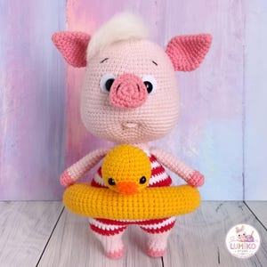 May include: A crocheted pig doll with light pink skin, a tuft of white hair, and large black eyes. The pig wears a red and white striped swimsuit and holds a yellow crocheted duck-shaped swim ring around its waist. A logo reads Lumiko Store.