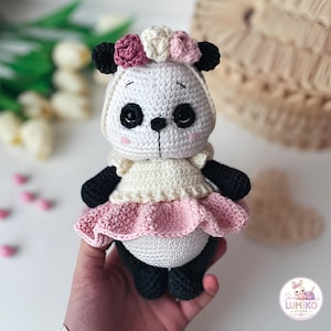 Könnte beinhalten: Eine handgefertigte Häkel-Panda-Puppe mit weiß-schwarzem Körper, die ein cremefarbenes Oberteil mit einem gerüschten rosa Rock trägt. Der Panda hat einen Blumenkranz aus gehäkelten Rosen in Rosa- und Weißtönen sowie schwarze Augen und rosa Wangen.