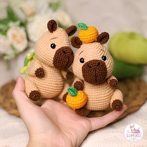 May include: Two crocheted capybara figures made of tan yarn with dark brown snouts, ears, and feet. One capybara has a small orange fruit with a green leaf on its head, and the other holds an identical orange fruit in its front paws.