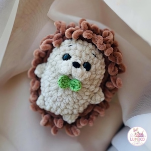 May include: A small, handmade crochet hedgehog plush toy featuring a cream-coloured body with a textured surface, dark brown curly yarn quills, two black bead eyes, a black nose, and a small green bow tie attached to the front of the chest.