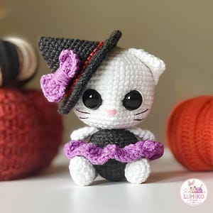 Può includere: Una bambola gatto all'uncinetto bianca con grandi occhi neri e un piccolo naso rosa. Il gatto indossa un cappello a punta nero con un fiocco viola e un vestito nero con una gonna a balze viola. Un piccolo logo nell'angolo riporta Lumiko Store.
