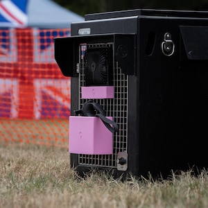 Puede incluir: Una jaula de metal negro para mascotas con un ventilador negro montado y dos cajas de almacenamiento de plástico rosa claro fijadas a la rejilla. Una caja contiene una correa negra con asa. La jaula tiene un cierre de metal plateado y un logo.