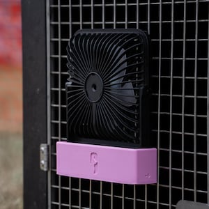Puede incluir: Un ventilador portátil negro con marco cuadrado y rejilla circular, montado sobre una base rectangular de color rosa claro. La base presenta una pequeña silueta de perro en relieve y un icono de huella en el lado derecho, sujeto a una rejilla metálica.