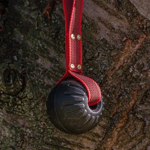 May include: A black spherical toy with a textured surface featuring molded ridges, attached to a red nylon strap. The strap is secured with two metallic rivets and forms a loop for holding, creating a durable design for active play and interaction.