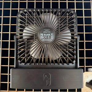 Può includere: Un ventilatore da scrivania portatile nero con telaio quadrato e un centro circolare contenente specifiche tecniche e simboli normativi. La base presenta un logo stilizzato a forma di testa di animale. Le pale sono visibili dietro la griglia.