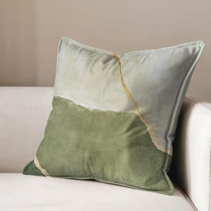 May include: A square throw pillow featuring a color-blocked design with a light sage green upper section and a darker olive green lower section. A thin, diagonal gold-colored line separates the two green tones, adding a metallic accent.