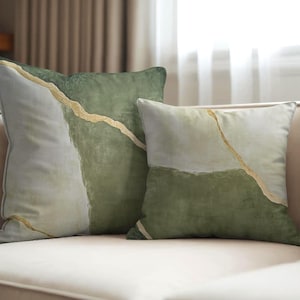 May include: Two decorative throw pillows with abstract designs. The pillows feature a color palette of green, white, and gold. The design includes a gold line across the pillow. The pillows are on a cream-colored sofa.