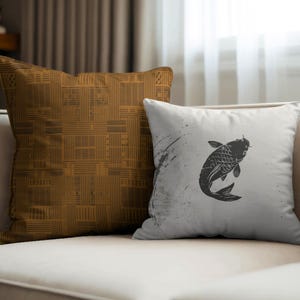 May include: Two decorative throw pillows on a cream-colored sofa. One pillow is a solid mustard color with a geometric pattern. The other pillow is white with a black koi fish illustration and abstract paint splatter.