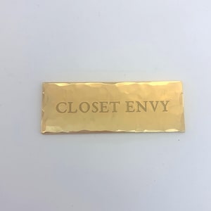 Könnte beinhalten: Rechteckige, goldfarbene Metallplakette mit dem Text "CLOSET ENVY" in der Mitte eingraviert. Die Plakette hat eine gehämmerten Textur und ist vor einem weißen Hintergrund platziert.