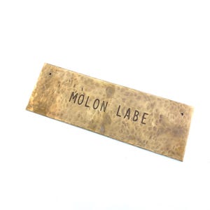 Könnte beinhalten: Eine rechteckige Messingplatte mit einer gehämmerten, strukturierten Oberfläche. Die Metallplatte trägt den in dunklen Großbuchstaben eingestanzten Text MOLON LABE. In der Nähe der beiden kürzeren Seitenränder befindet sich jeweils ein kleines Loch.