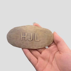 Könnte beinhalten: Ovales Holzschild mit den Initialen "HJL" in einer Serifenschrift graviert. Das Schild hat eine natürliche Holzmaserung. Das Schild wird in einer Hand gehalten.