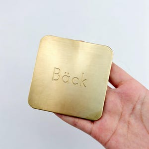 Könnte beinhalten: Ein quadratischer Untersetzer aus gebürstetem goldfarbenem Metall mit abgerundeten Ecken. Das Wort Bäck ist mittig in einer klaren, serifenlosen Schrift eingraviert. Die Oberfläche hat eine feine horizontale Maserung, die das Licht reflektiert.