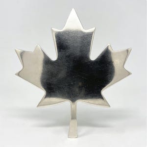 Peut inclure: Un objet en métal argenté poli en forme de feuille d'érable. L'article présente une surface lisse et réfléchissante avec des lobes pointus distincts et une tige centrale à la base, offrant une silhouette plate et symétrique sur un fond uni.
