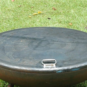 May include: A round, metal fire pit with a lid. The lid has a handle and is closed. The fire pit is sitting on a grassy surface.