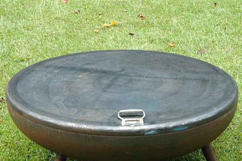 May include: A round, metal fire pit with a lid. The lid has a handle and is closed. The fire pit is sitting on a grassy surface.