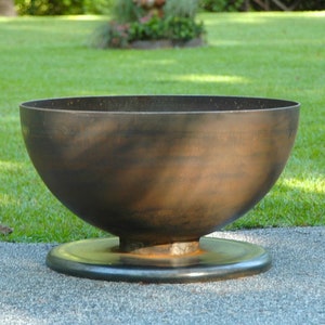 May include: A large, round, metal fire pit with a wide, shallow bowl and a raised base. The fire pit is made of dark brown metal and has a rustic, weathered look.