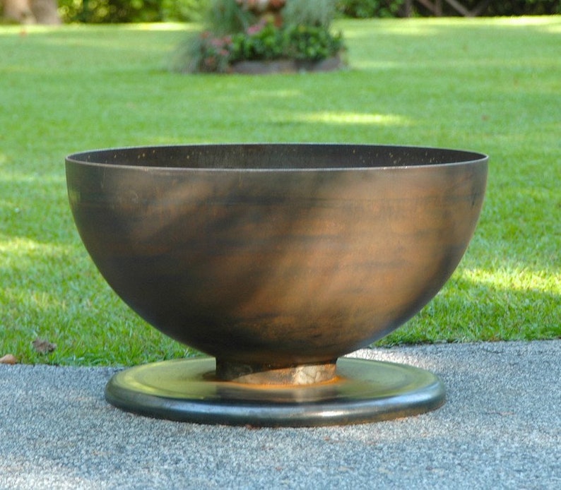 May include: A large, round, metal fire pit with a wide, shallow bowl and a raised base. The fire pit is made of dark brown metal and has a rustic, weathered look.