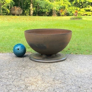 May include: A large, rusty metal bowl sits on a round base. The bowl is about the size of a bowling ball, which is sitting next to it on the ground.