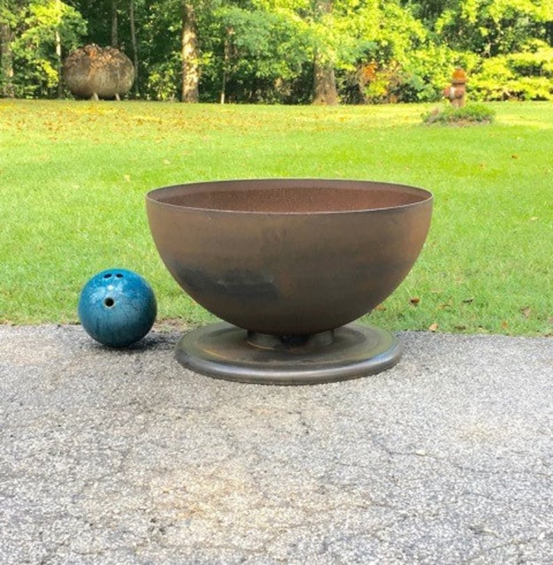 May include: A large, rusty metal bowl sits on a round base. The bowl is about the size of a bowling ball, which is sitting next to it on the ground.
