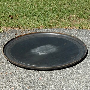 May include: A large, round, black metal plate with a slightly raised edge. The plate has a worn, rustic look and is sitting on a gray gravel surface.