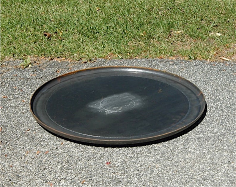 May include: A large, round, black metal plate with a slightly raised edge. The plate has a worn, rustic look and is sitting on a gray gravel surface.