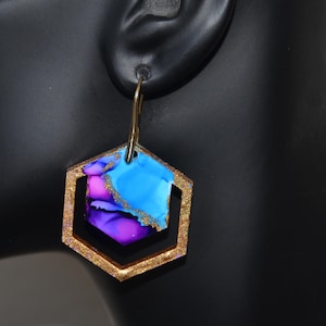 May include: A gold-colored, hexagonal earring with a colorful, abstract design. The inner hexagon features blue, purple, and gold accents. The earring hangs from a gold-colored hook, showcasing a modern, artistic style.
