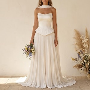 May include: A strapless ivory wedding dress with a fitted bodice and pleated skirt. The dress features a sweetheart neckline and a peplum waist. A matching cape drapes over the shoulders. The bride holds a bouquet of wildflowers.