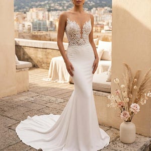 Puede incluir: Un vestido de novia blanco con silueta de sirena. El corpiño presenta detalles de encaje intrincados y un escote pronunciado. El vestido tiene una larga cola y se muestra al aire libre.