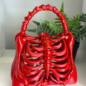 May include: A bright red handbag shaped like a human rib cage. The bag features a rigid, skeletal structure with a curved handle formed by connected bone segments. The surface has a smooth, glossy finish, and the design mimics the anatomy of a torso.