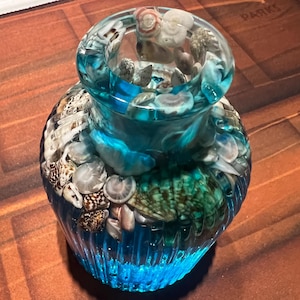 Könnte beinhalten: Eine dekorative Glasvase gefüllt mit Muscheln und kleinen Steinen. Die Vase hat einen türkisfarbenen Farbton und ist mit verschiedenen Muscheln verziert. Der Boden der Vase hat ein strukturiertes, geripptes Design.