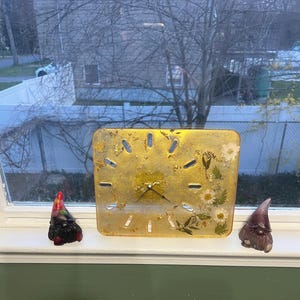 Könnte beinhalten: Eine quadratische, goldfarbene Uhr mit schwarzen Zeigern und eingebetteten getrockneten Blumen. Zwei kleine Zwergfiguren sitzen auf beiden Seiten der Uhr. Die Uhr steht auf einer Fensterbank mit Blick auf ein Wohngebiet.