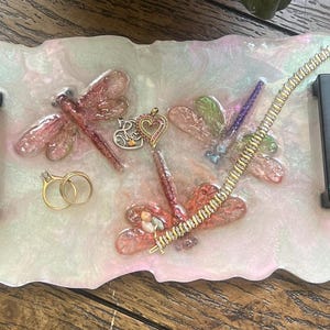 May include: A decorative tray with a pearlescent finish, featuring embedded dragonfly designs in pink, green, and purple. Gold rings, a bracelet, and a heart-shaped pendant are also on the tray. Black handles are attached to the sides.