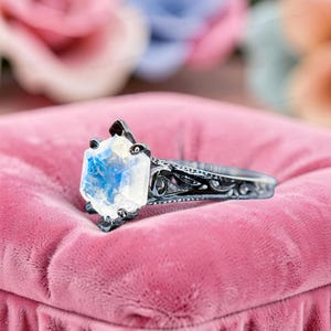 May include: A ring featuring a hexagonal moonstone with a blue iridescent sheen, set in a dark metal band with intricate, cut-out scrollwork patterns along the sides. The gemstone is held in place by six small prongs extending from the metal setting.