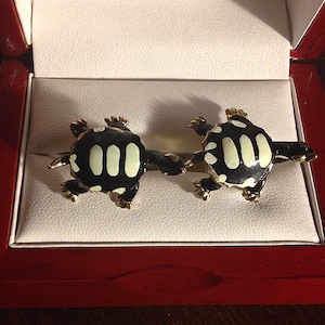 May include: A pair of black and white turtle cufflinks in a red velvet box. The turtles are made of metal and have a shiny finish.