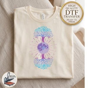 May include: A cream-colored t-shirt featuring a central graphic of a moon positioned between two mirrored tree designs. The trees and moon are rendered in shades of blue and purple with radiating lines. A gold circular badge reads HIGH QUALITY DTF TRANSFER.