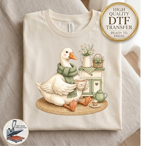 May include: A cream-colored t-shirt featuring a graphic of a white goose wearing a green knit scarf, holding a floral teacup. The scene includes a small cabinet, a pitcher of daisies, a basket, a green teapot, and a cupcake on a woven rug.