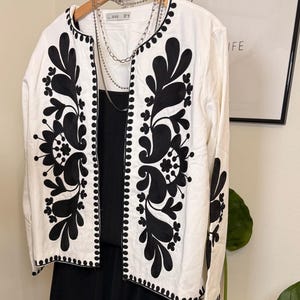 May include: A white jacket with black floral embroidery and a black dress. The jacket has a round collar and long sleeves. Silver necklaces hang from the jacket. The jacket is hanging on a wooden hanger.