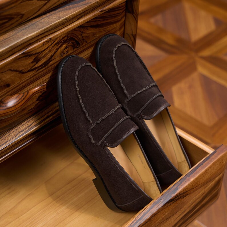 May include: A pair of dark brown suede loafers featuring a scalloped edge detail along the vamp and a black sole. The shoes have a smooth tan interior lining and a low heel, showcasing a classic slip-on design with clean lines and a soft texture.