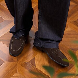 May include: A pair of dark brown suede loafers with a scalloped edge detail along the vamp, worn with dark charcoal gray denim trousers featuring visible white stitching along the hemline and straight-leg cut.