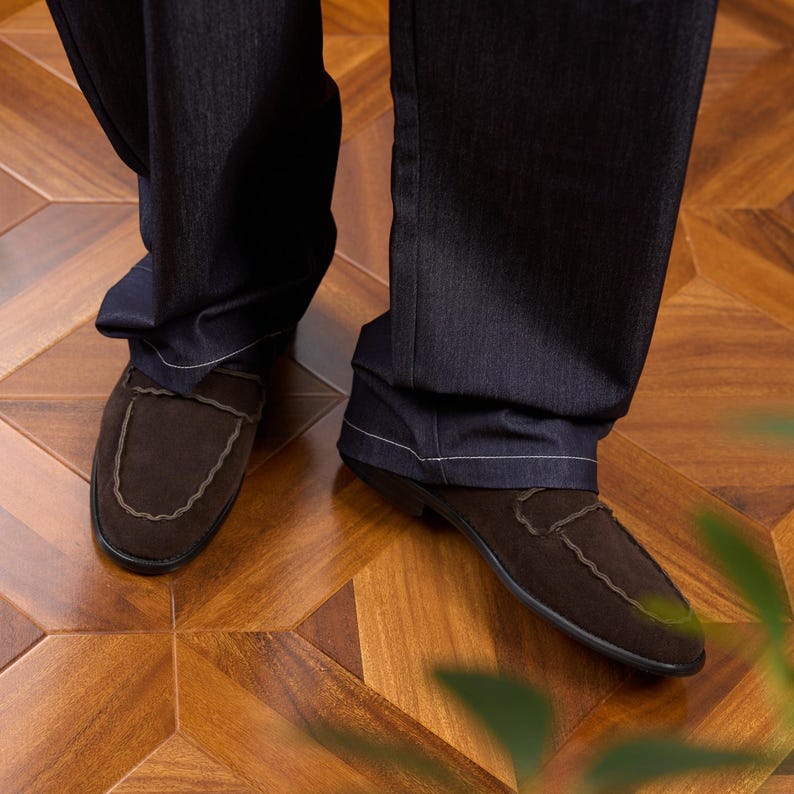 May include: A pair of dark brown suede loafers with a scalloped edge detail along the vamp, worn with dark charcoal gray denim trousers featuring visible white stitching along the hemline and straight-leg cut.