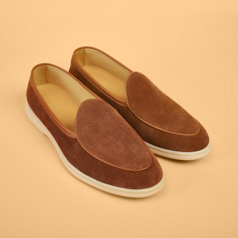 Op de afbeelding: Een paar bruine su&egrave;de loafers met een ronde neus en een witte zool. De schoenen hebben een klassiek design met een gladde, getextureerde uitstraling. De binnenkant is lichtbruin.