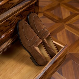 May include: A pair of brown suede loafers featuring a moccasin-style stitched toe seam and a low heel. The shoes have a smooth tan interior lining and a dark brown sole, presented with a clean finish and visible texture on the suede material.